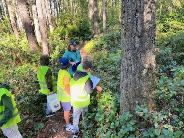 A Thurston County employee leads an educational plant walk with students through a wooded area. Students are wearing high visibility vests and are holding clipboards. 