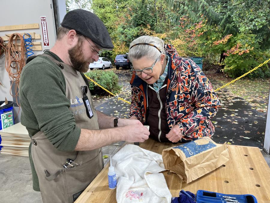 Photo taken of attendees at a recent Fix-It Fair. A man holds a newly repaired soapdish, showing to an attendee near a workbench.
