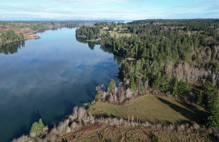 aerial photo of an open grassy field in the middle of a forested area along a waterbody