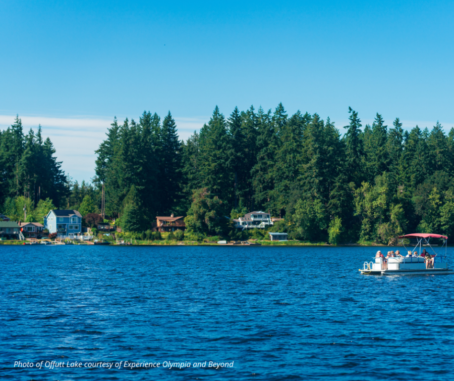 Photo of Offutt Lake in Thurston County on a sunny day. The dark blue lake is edged by tall evergreen trees and a few houses dotting the shoreline. In the mid-distance is a small boat.