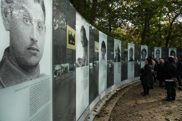 Im linken Teil des Fotos sind Wandtafeln aufgestellt, auf denen Fotos und Informationstexte abgebildet sind. Im rechten Teil des Fotos stehen Menschen, die sich die Informationstafeln anschauen. Im oberen Teil des Fotos sind Bäume zu sehen.