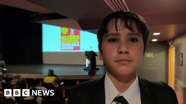 A young boy looking at the camera. He has dark hair that covers his forehead. He has a dark blazer on, with a white shirt and stripped tie. He is in a school hall