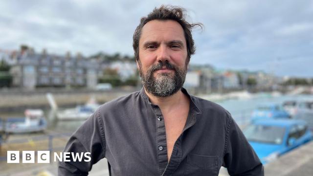 A man with dark brown air and a greying beard looking at the camera. He is wearing a dark shirt with the top three buttons undone. You can see St Peter Port seafront in the back.