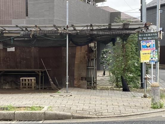 A view of a partially closed building with a wooden structure and a broom leaning against it. A sign with contact information is visible, alongside some greenery and a stone curb. The scene appears to be in an urban setting.