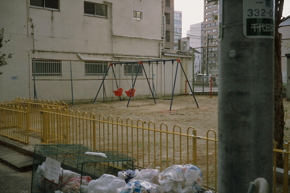街なかにある小さな公園の写真。画面の中央にはベージュ色の地面がひろがっていて2組のブランコがある。画面の手前側には公園の出入り口があり、公園は黄色い柵で囲まれている。出入り口の右側は収集に出されたゴミ袋、さらに右端には電信柱が見えている。公園の左奥は古そうなビルのベージュ色の壁があり、画面中央から右にかけて、公園の向こうには駐車場や高層ビルが見えている。
