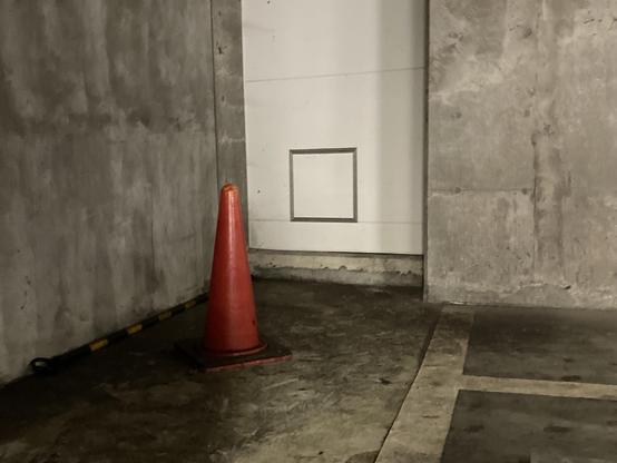 A bright orange traffic cone stands in a dimly lit concrete area, next to a white wall with a square inset. The floor shows signs of wear, and a yellow and black striped barrier is visible along the lower edge.
