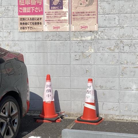 The image shows a grey car parked near a wall with peeling paint. Two traffic cones with the words "封鎖中" (meaning "closed" in Japanese) are placed on the ground. Above them, there are posters with instructions related to