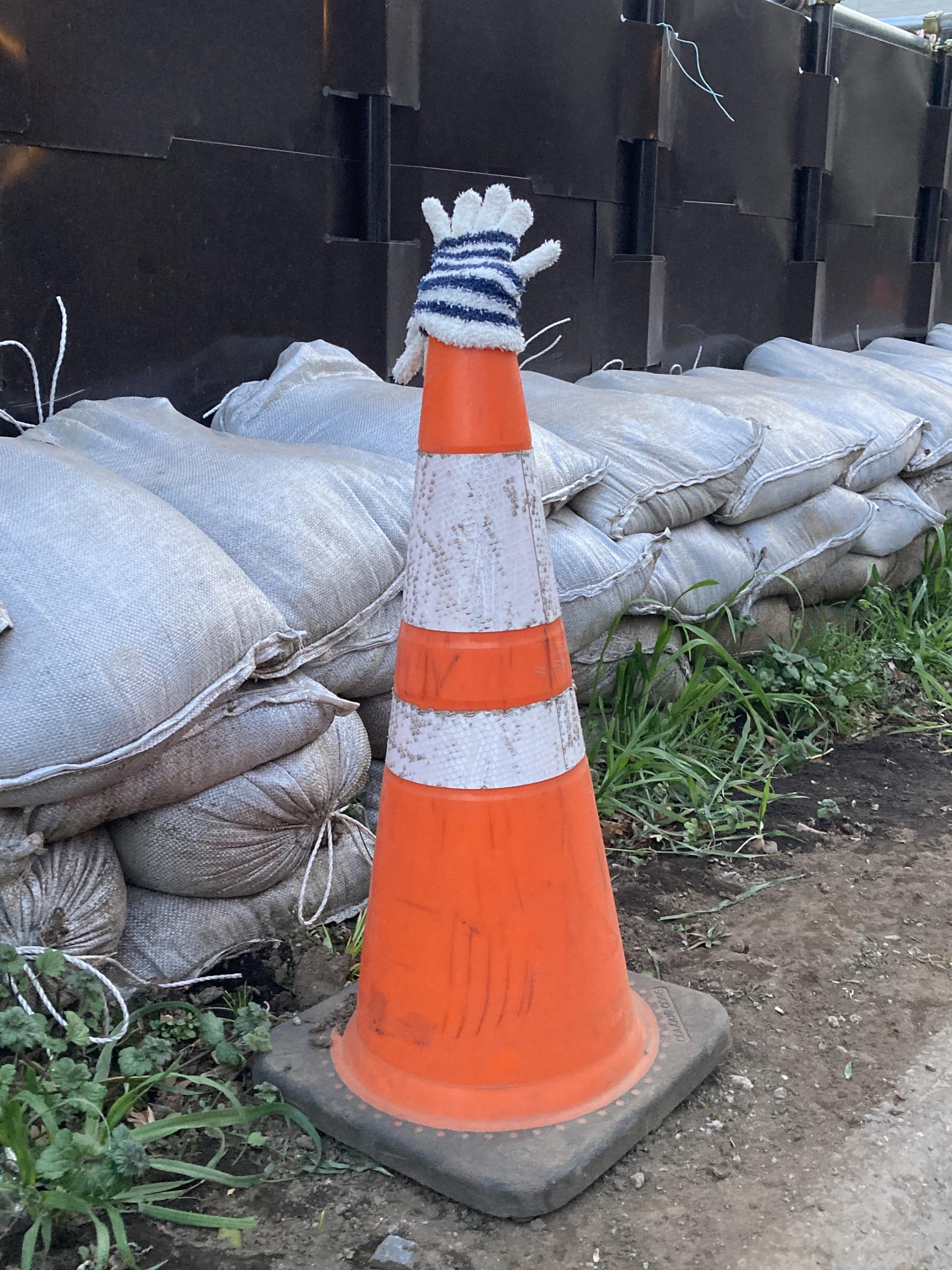 An orange traffic cone is placed next to a stack of sandbags, with a white and blue striped glove resting on top of the cone. Grass and dirt are visible in the surrounding area.