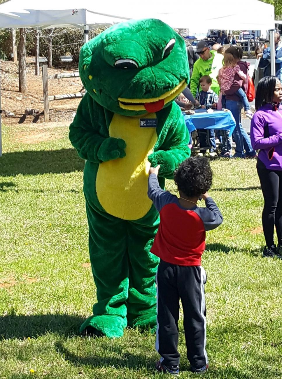 frog mascot stands with child on open play field with tents and trees in background