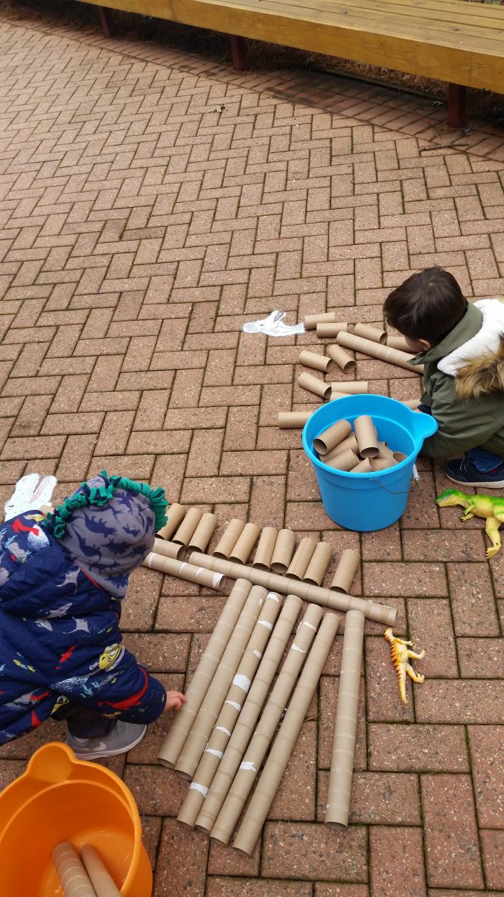 Preschool aged kids making a dinosaur skeleton out of cardboard tubes on the brick patio in front of the Education Center