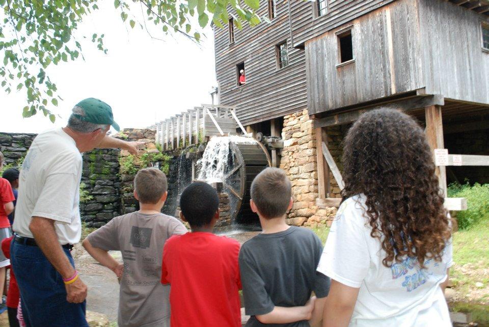 Group of school children stand outside old wooden mill building and waterfall