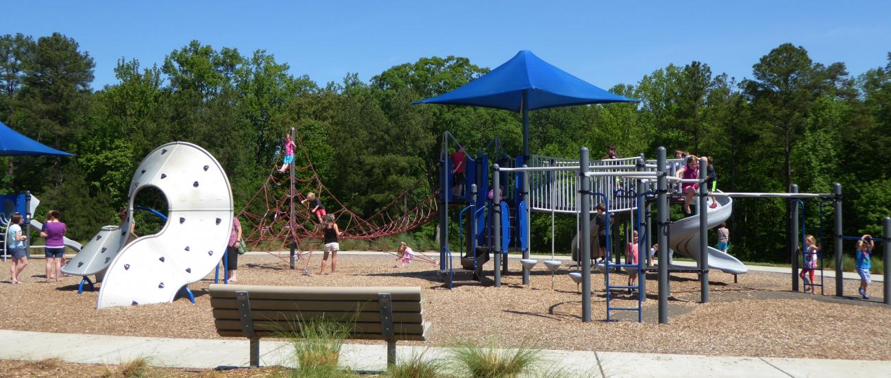Children playing on the playground under sunny skies while adults supervise.