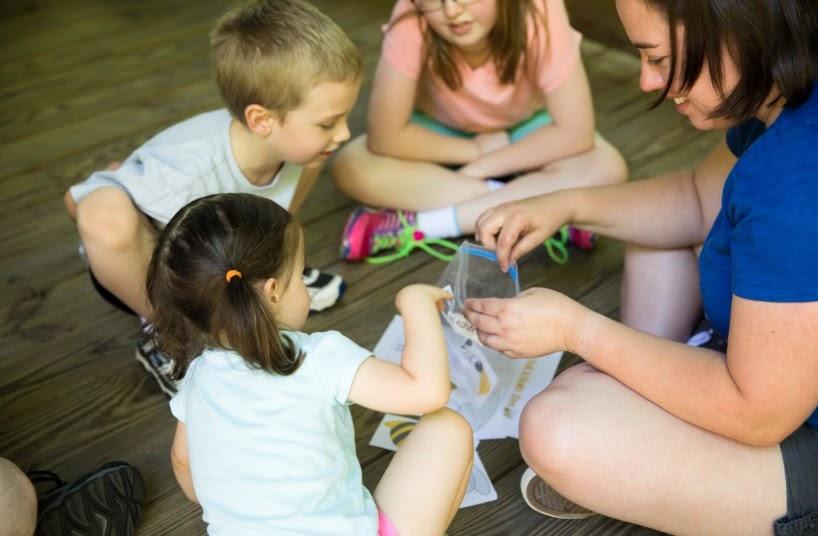 woman works with three children on crafts on wooden floor of pavilion