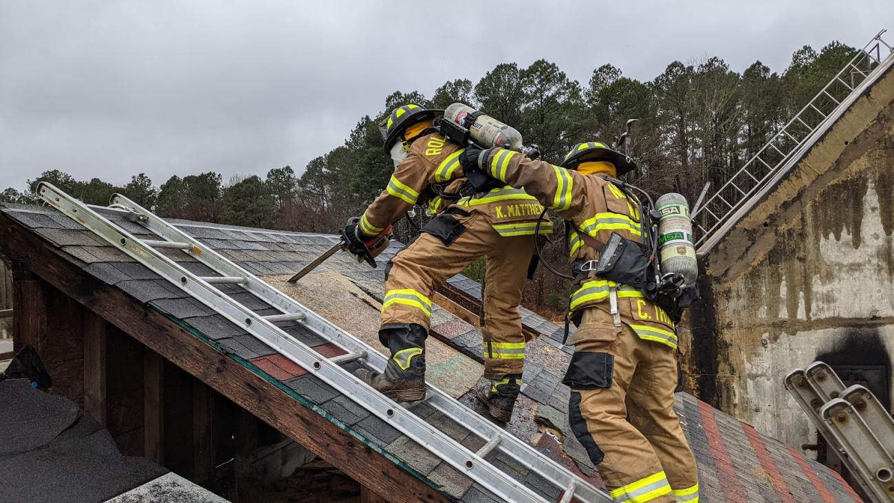 Two Fire Academy trainees scale a roof and use a saw