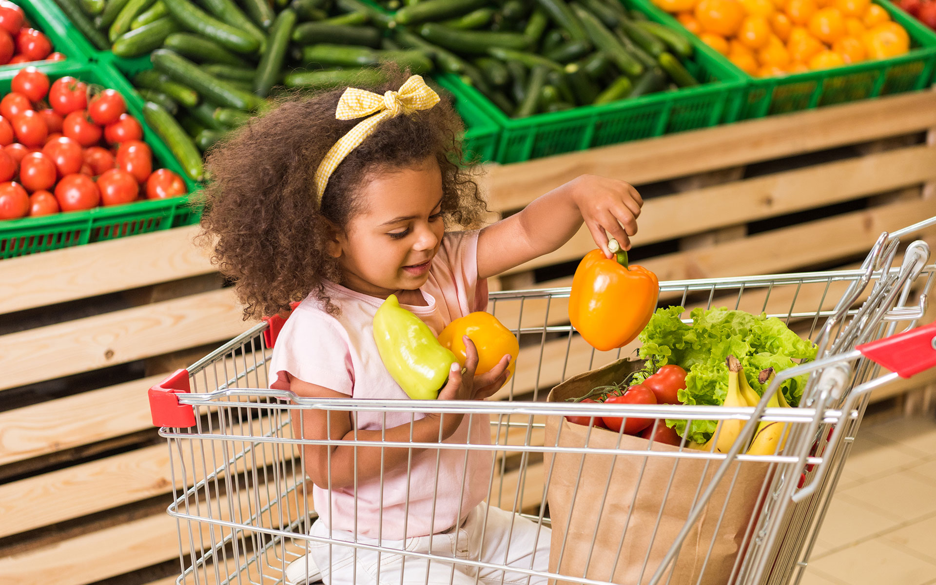 A child in a grocery cart holding three yellow bell peppers.