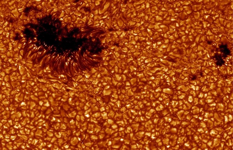 A view of an irregular-shaped sunspot and granules on the Sun's surface. The sunspot is dark, and the Sun's surface is orange with a granulated texture.